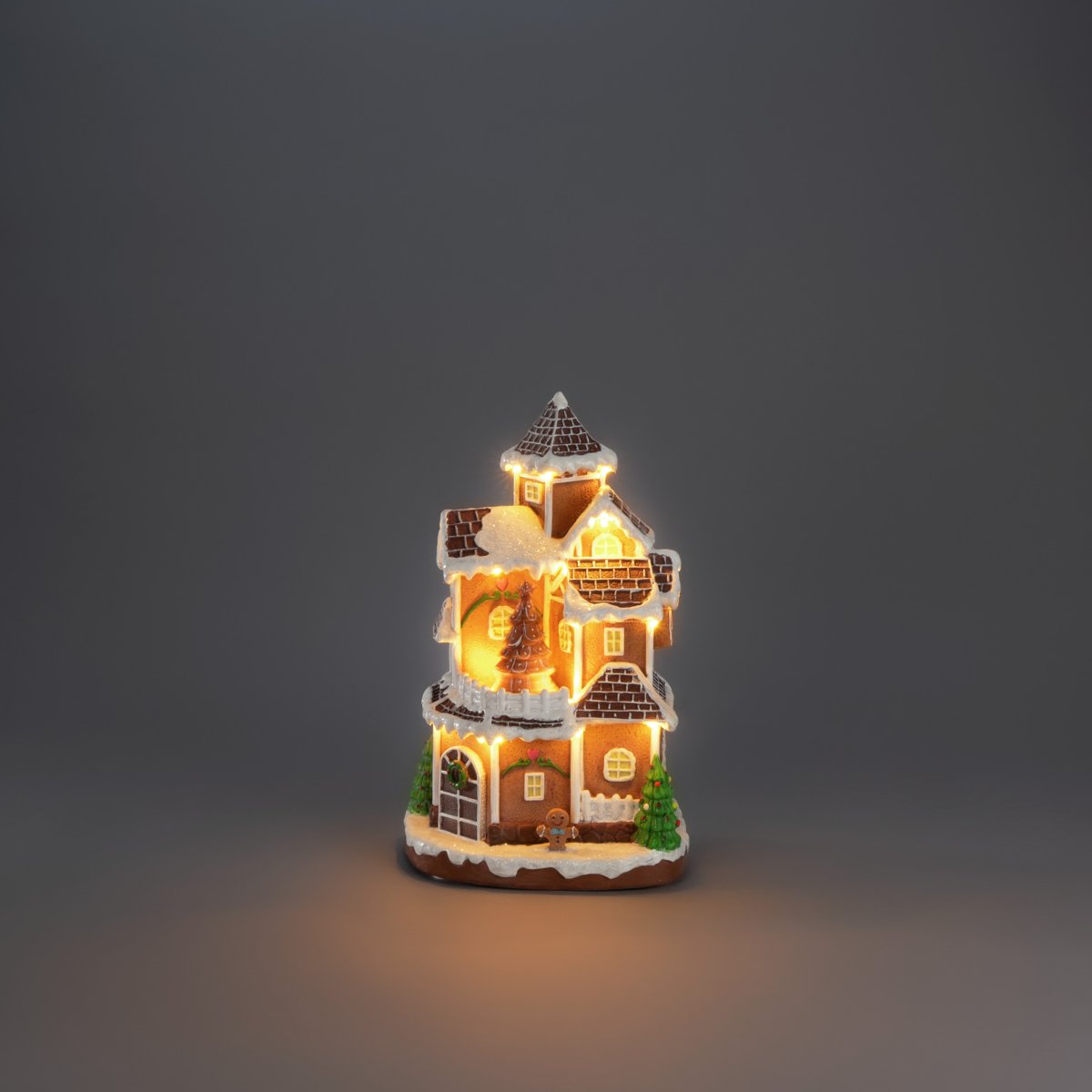 Pepperkakeby 30cm Brun Fra Konstsmide - Det detaljerte huset med sn&oslash;dekt tak, skinnende vinduer og dekorert grantre sprer en varm og innbydende gl&oslash;d i rommet.