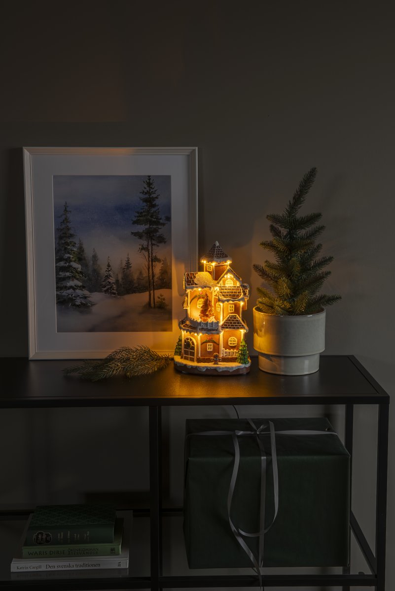 Pepperkakeby 30cm Brun Fra Konstsmide - Det detaljerte huset med sn&oslash;dekt tak, skinnende vinduer og dekorert grantre sprer en varm og innbydende gl&oslash;d i rommet.