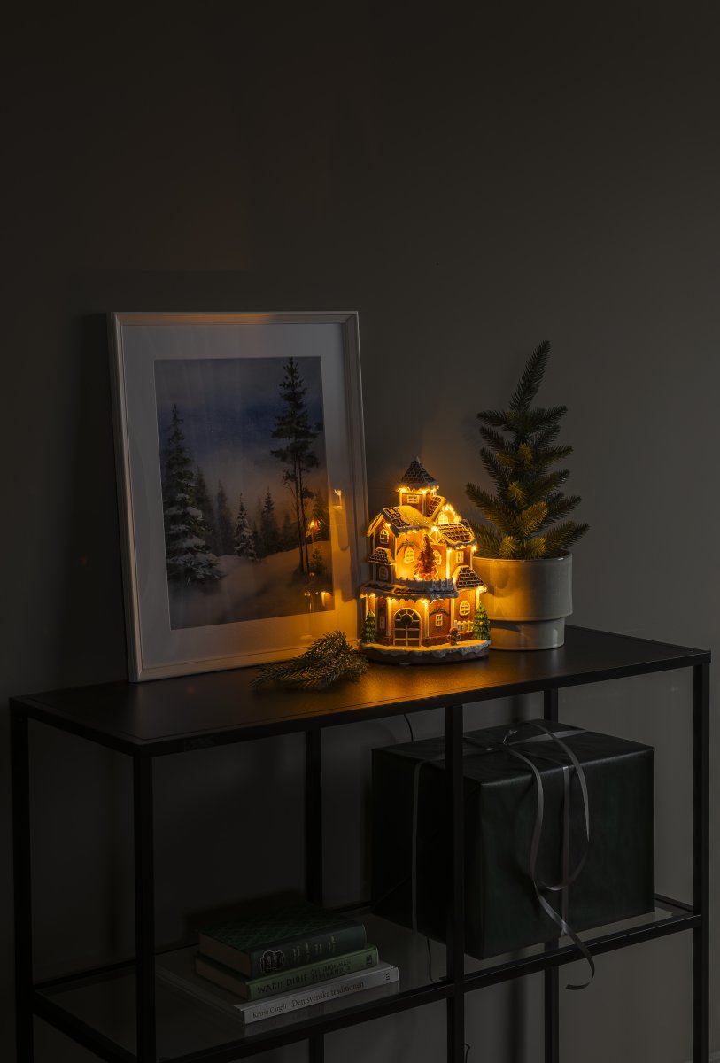 Pepperkakeby 30cm Brun Fra Konstsmide - Det detaljerte huset med sn&oslash;dekt tak, skinnende vinduer og dekorert grantre sprer en varm og innbydende gl&oslash;d i rommet.