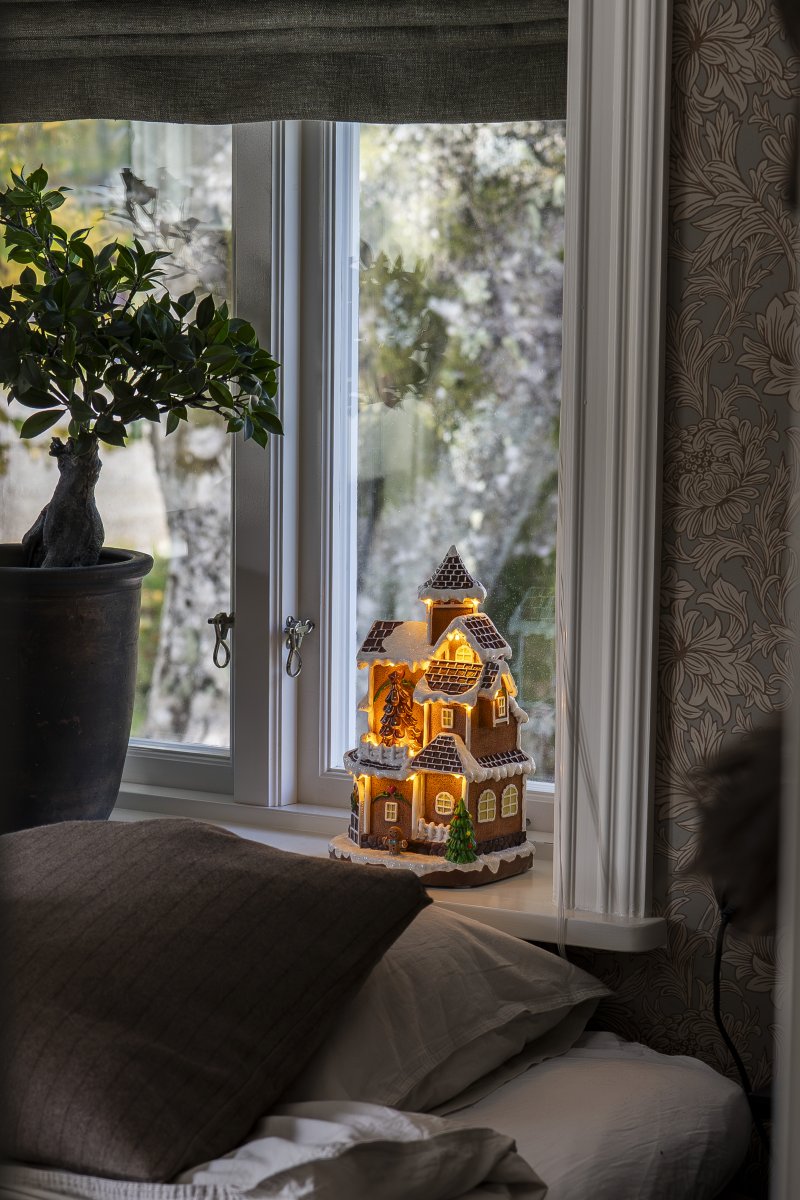 Pepperkakeby 30cm Brun Fra Konstsmide - Det detaljerte huset med sn&oslash;dekt tak, skinnende vinduer og dekorert grantre sprer en varm og innbydende gl&oslash;d i rommet.