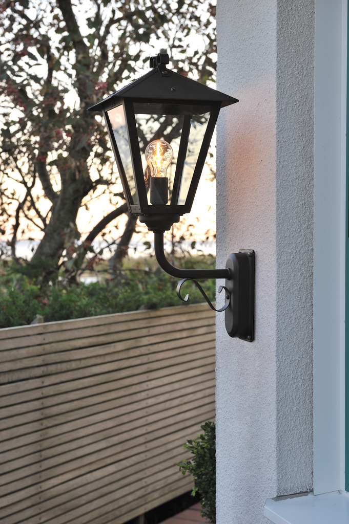 Benu Svart utendørslampe Fra Konstsmide - Lampens klassiske lyktform og robuste materiale gj&oslash;r den til et perfekt valg for de som &oslash;nsker &aring; skape en innbydende atmosf&aelig;re ved innganger eller lange korridorer.