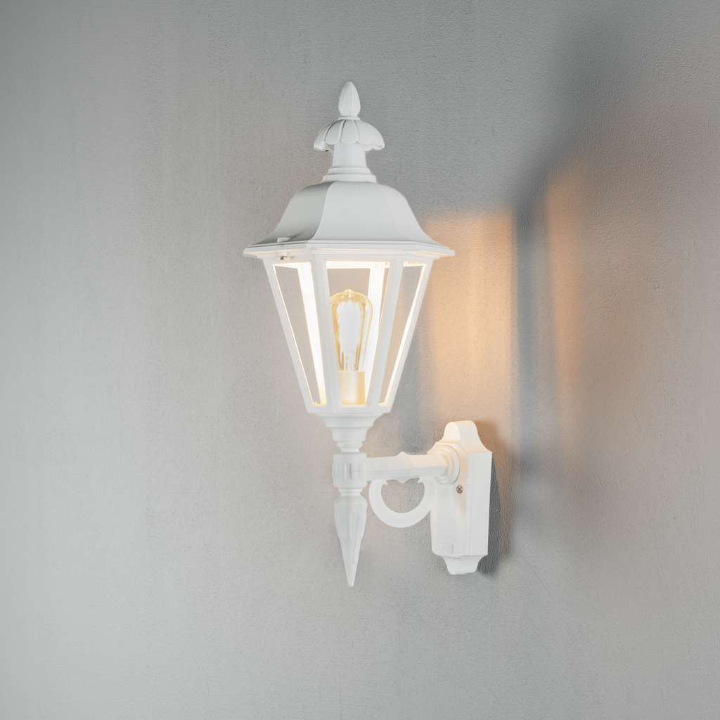 Pallas Hvit utendørslampe Fra Konstsmide - Med sitt lanterneformede design og klare glassfelt sprer lampen et varmt og innbydende lys som er perfekt for innganger, uteplasser og gangveier.