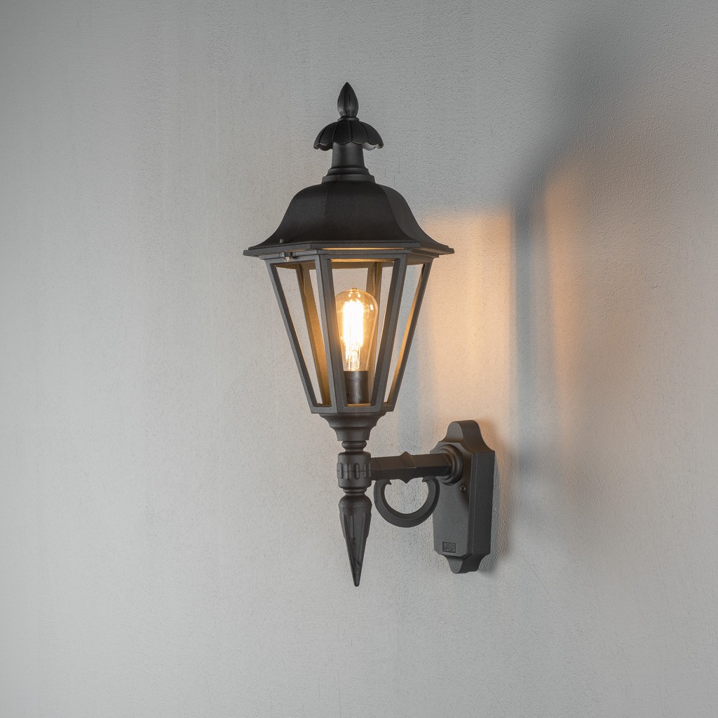 Pallas Svart utendørslampe Fra Konstsmide - Med sitt lanterneformede design og klare glassfelt sprer lampen et varmt og innbydende lys som er perfekt for innganger, uteplasser og gangveier.