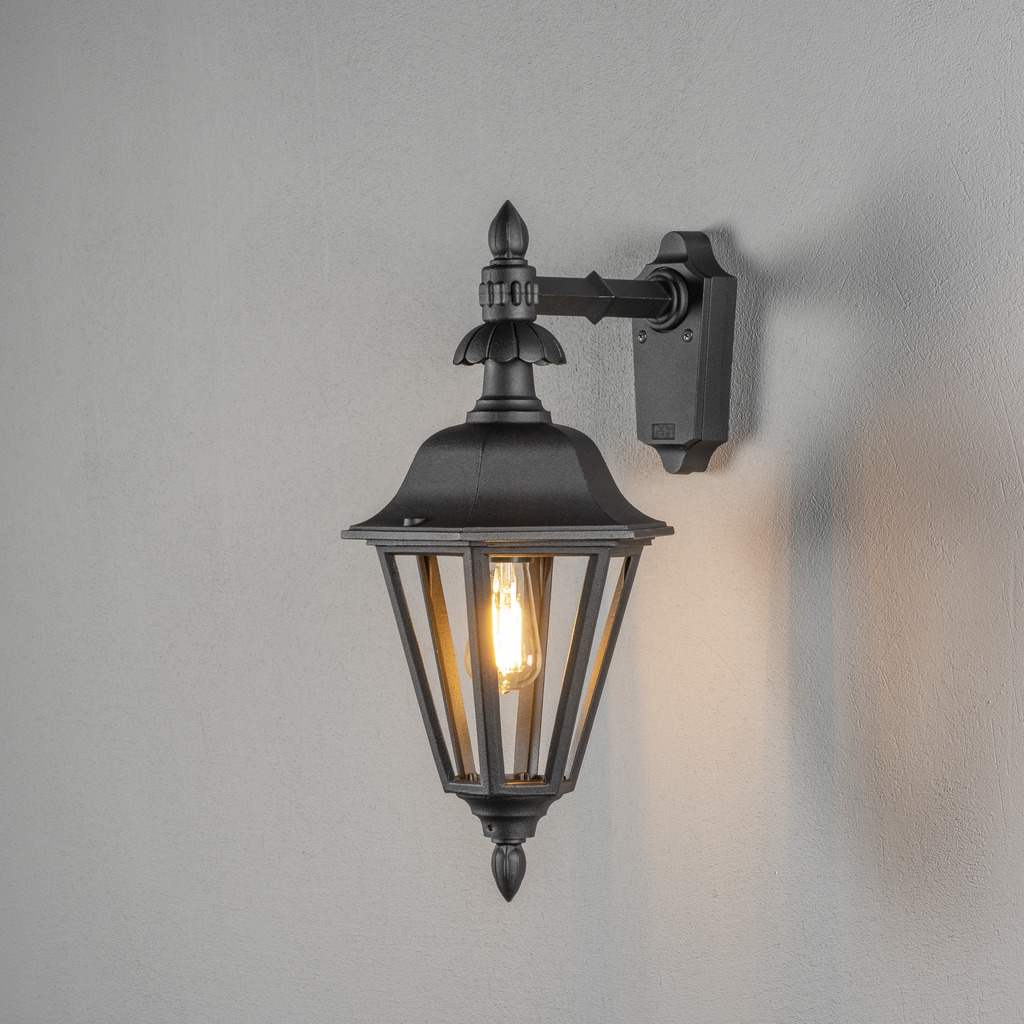 Pallas Svart utendørslampe Fra Konstsmide - Med sitt lanterneformede design og klare glassfelt sprer lampen et varmt og innbydende lys som er perfekt for innganger, uteplasser og gangveier.