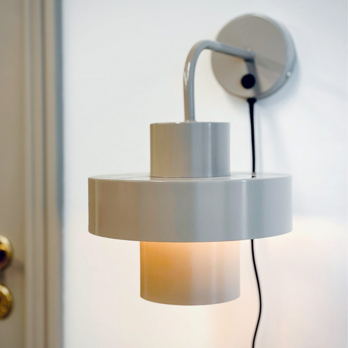 Stoke Ø15 Varm grå vegglampe Fra Dyberg Larsen - Den er laget av metall med en blank overflate som fremhever de sylindriske formene på lampen og skaper en balansert kontrast med andre materialer som stein, tre.