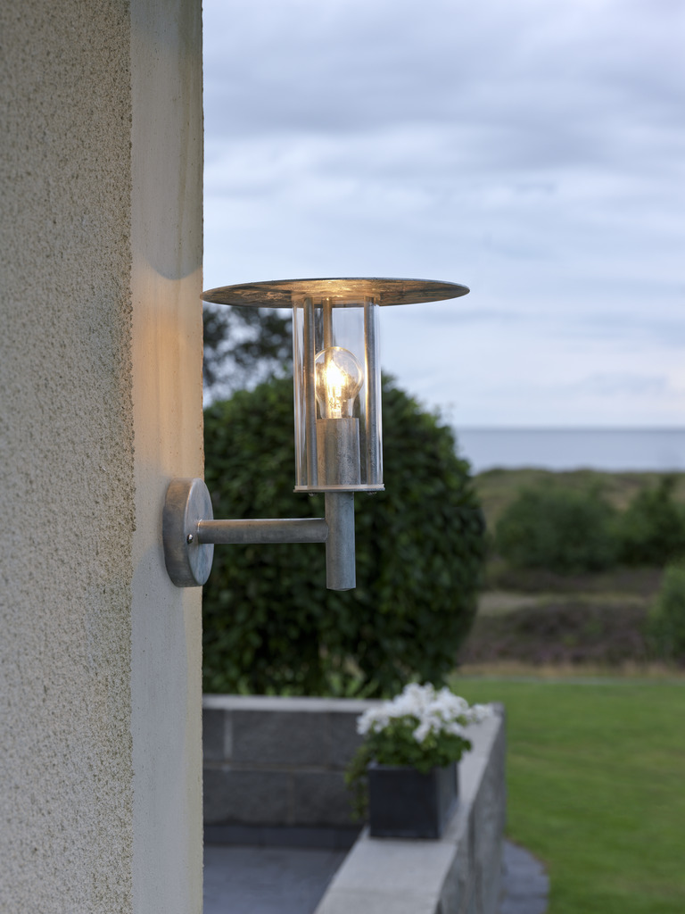 Mode Galvanisert stål utendørslampe Fra Konstsmide - Med sitt minimalistiske design og sylindriske glassbeholder sprer lampen et klart og behagelig lys som lyser opp overbygde innganger og uteplasser.