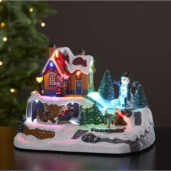 Winterville Flerfarget Fra Star Trading - En nydelig juledekorasjon med en sn&oslash;mann som kikker ut over sk&oslash;ytebarna og julenissen med slede og reinsdyr.