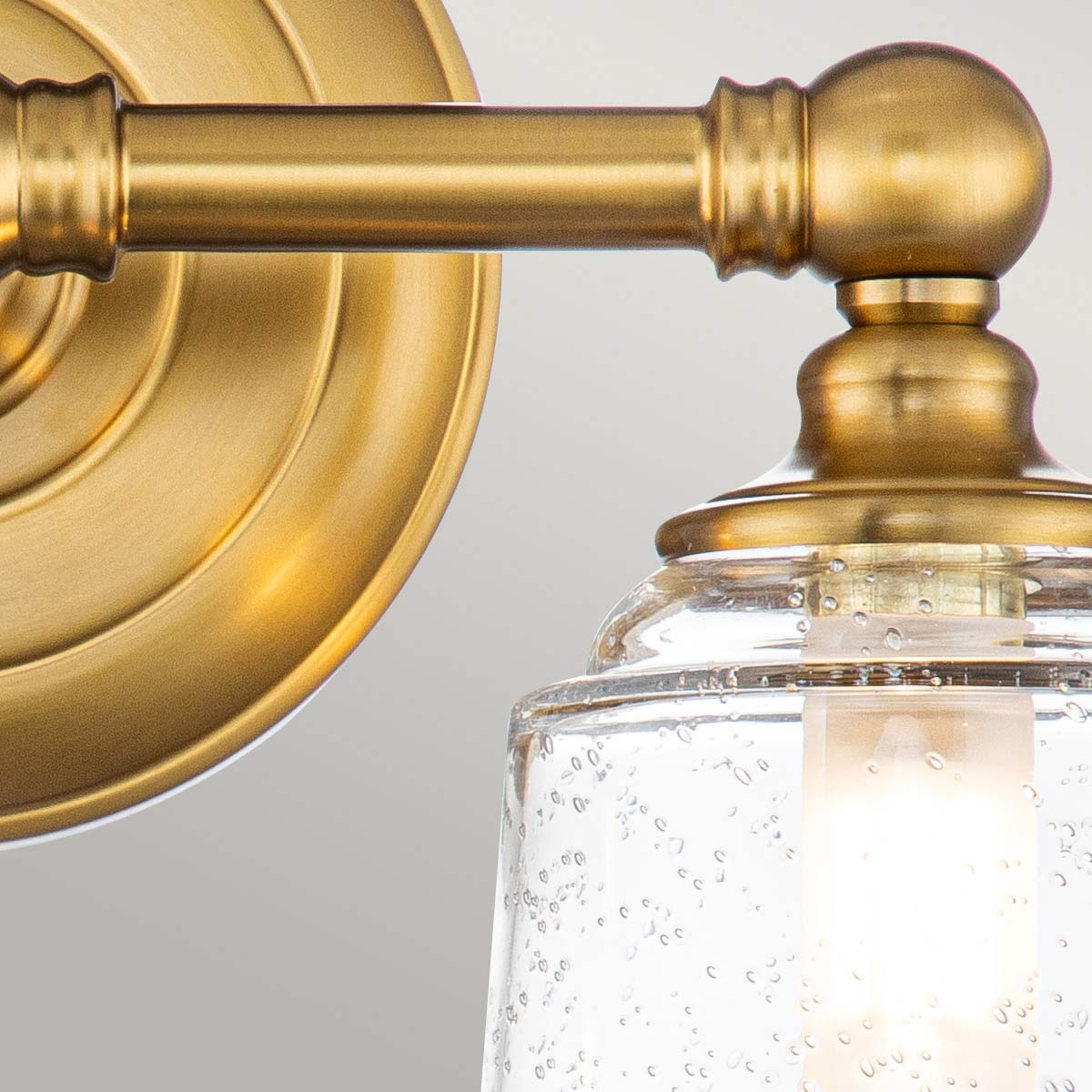 Huguenot Lake 33cm Messing baderomslampe Fra Elstead Lighting - De elegante sirkulære detaljene og håndkuttede brakettene gir en følelse av tradisjonelt håndverk, mens det klare, strukturerte glasset skaper et mykt og innbydende lys.